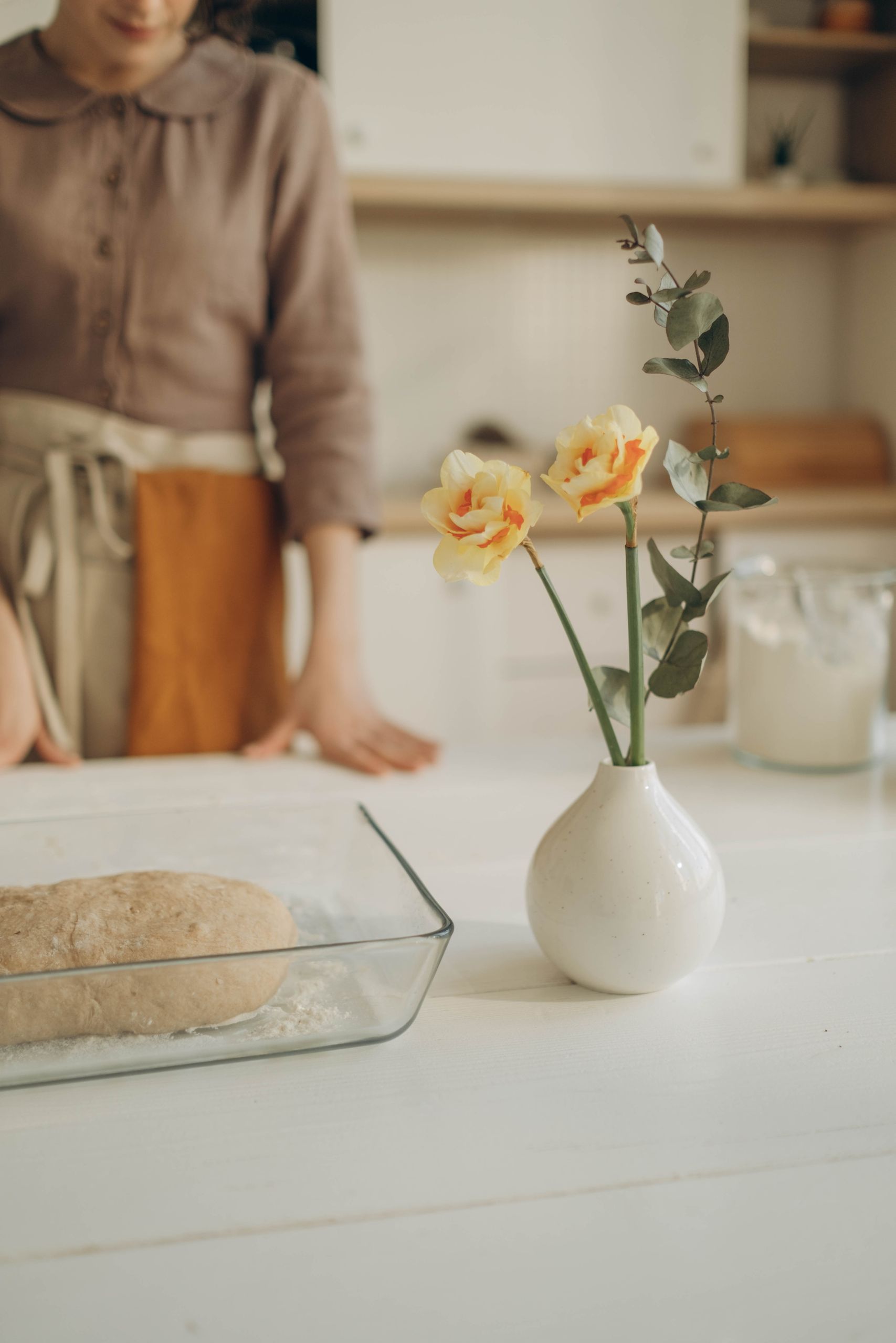 A cozy kitchen setting with rising dough in a glass tray and vase with flowers.