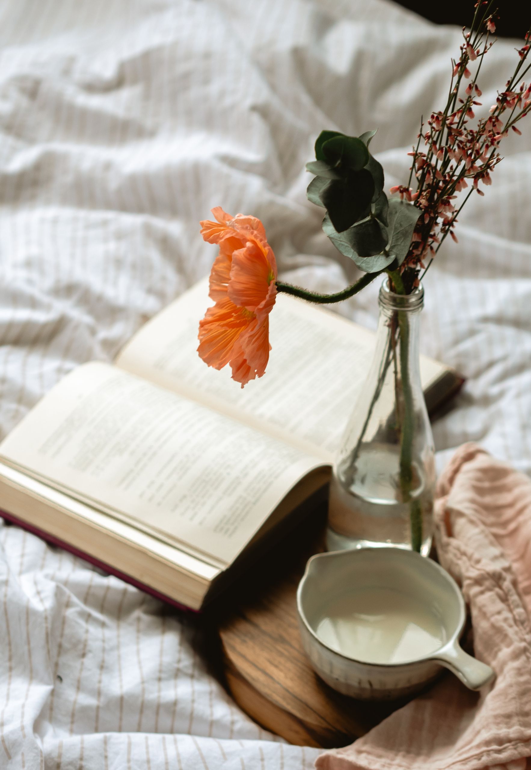 A warm and inviting setup with a book, flower, and milk on bed linen, perfect for relaxation.
