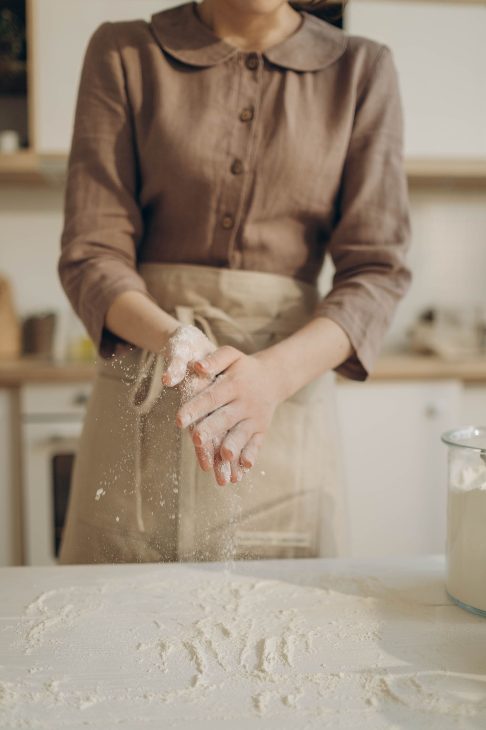 A woman claps hands over a table covered with flour in a cozy kitchen setting.